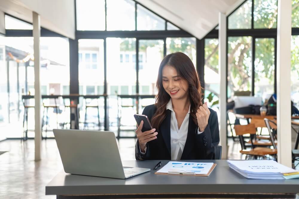 Woman sitting at a desk in front of a laptop, celebrating successfully discovering how to sell more policies and become a top-tier insurance agent.