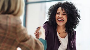 Smiling woman reaches out her hand to shake hands with an interviewer, seen from behind.