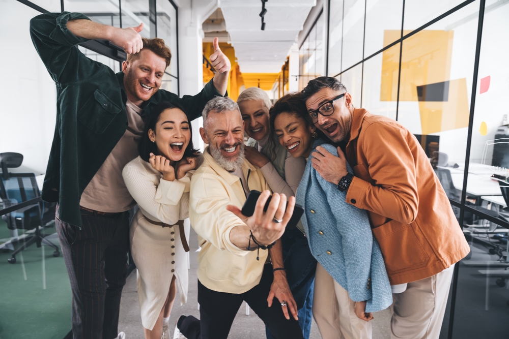 Diverse coworkers taking a group selfie, highlighting a multigenerational client base through teamwork and inclusion.