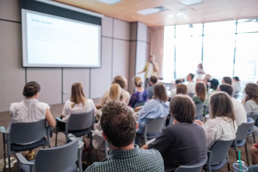 Attendees sit attentively as speaker presents during business conference. Bright, modern meeting room enhances learning experience while promoting collaboration and focus among participants. Insurance agent professional growth concept.
