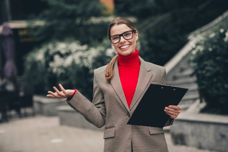 An insurance agent in fall-inspired attire holding a clipboard, presenting a confident and cheerful expression against an urban outdoor background, ready to use fall strategies for insurance agents.