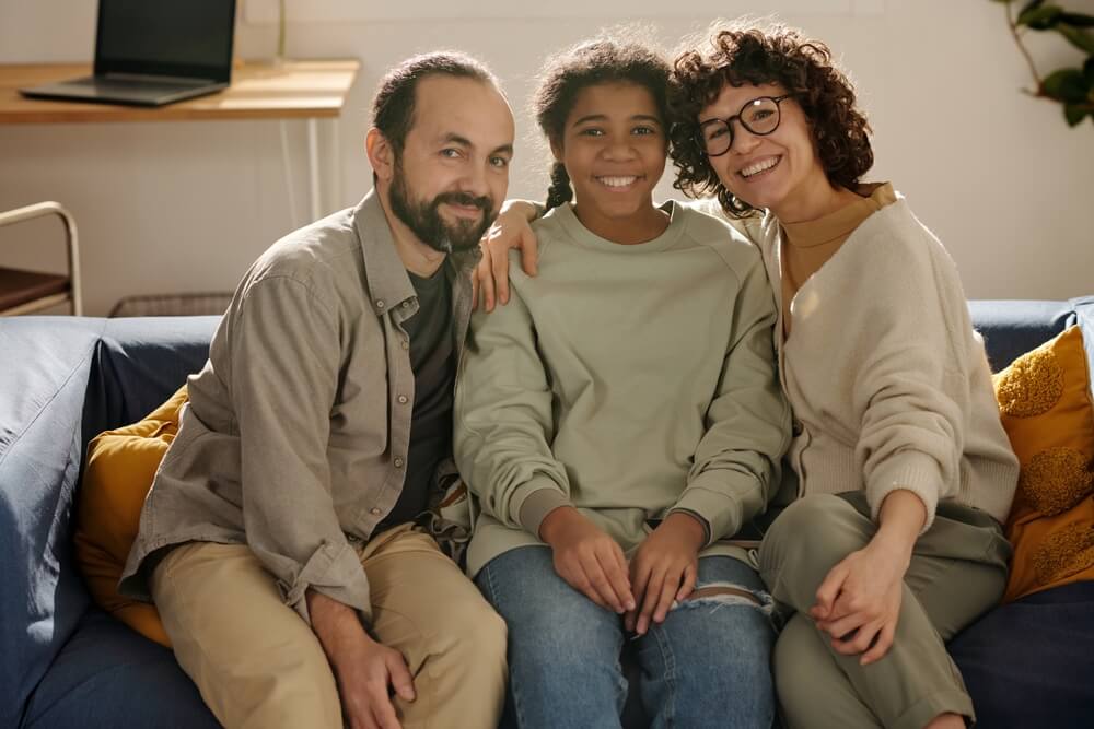 Portrait of happy parents sitting together with their adopted daughter on sofa and smiling at camera.