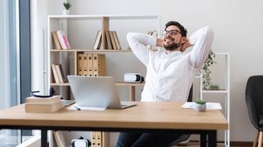 Young adult white male wearing glasses and a white shirt sitting at a desk smiling during an office break, demonstrating relaxation and independent insurance agents work-life balance.