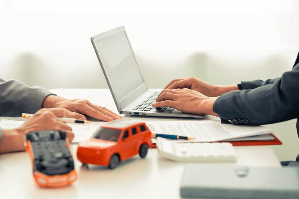 Independent agent discussing auto insurance coverage with client at desk, reviewing documents with laptop and car models visible