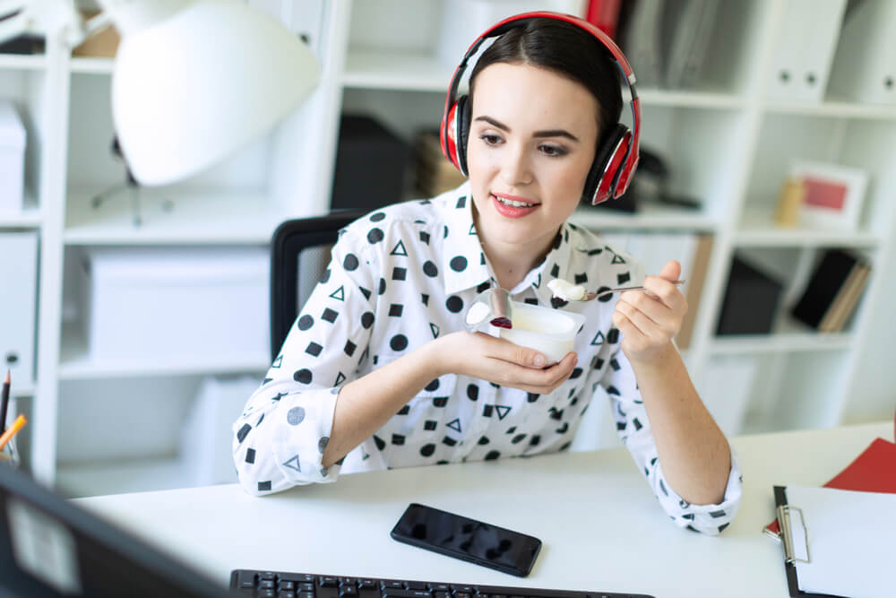 An independent insurance agent wearing headphones eats yogurt while working at a desk with documents and a smartphone in a modern office.