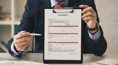 A professionally dressed insurance agent in a suit holds up a clipboard displaying an insurance form while pointing at it with a pen, representing insurance services and client documentation; the scene suggests professionalism, policy management, and CRM certification or training in customer relationship management within the insurance industry.