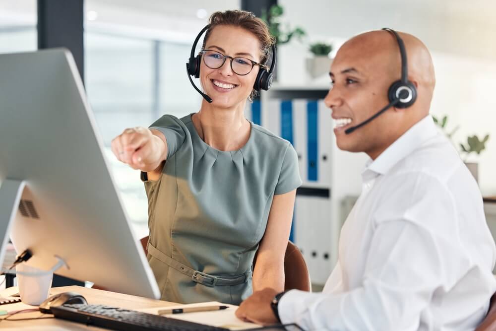 A customer service or insurance support team in a modern office, wearing headsets and collaborating at a computer, with one colleague pointing at the screen—suggesting CRM system training, teamwork, and CRM certification in a client-focused environment.